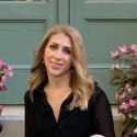 photo of Afsaneh Essatyar smiling long blond hair, dark eyes, wearing black v-neck blouse with pink flowers and green door in background