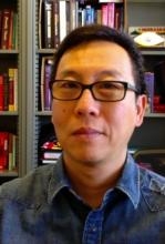 photo of Karl Kwan short brown hair, black glasses, blue shirt with bookshelf in the background