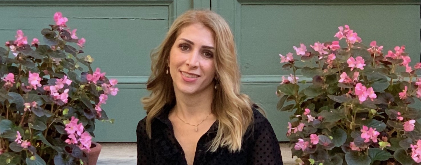 photo of Afsaneh Essatyar smiling long blond hair, dark eyes, wearing black v-neck blouse with pink flowers and green door in background