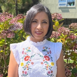 photo of Yolanda Gamboa smiling, dark medium length hair, dark eyes, short sleeved white blouse with colorful embroidered flowers. Background is outside with flowers behind Yolanda.