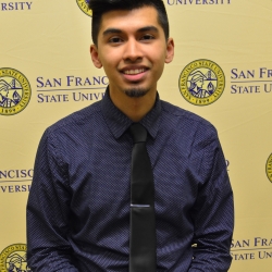 photo of Leo Cruz smiling short dark hair, dark eyes, light brown skins with short beard and mustache, wearing dark blue collared shirt with dark tie. The background is gold with SF State logos